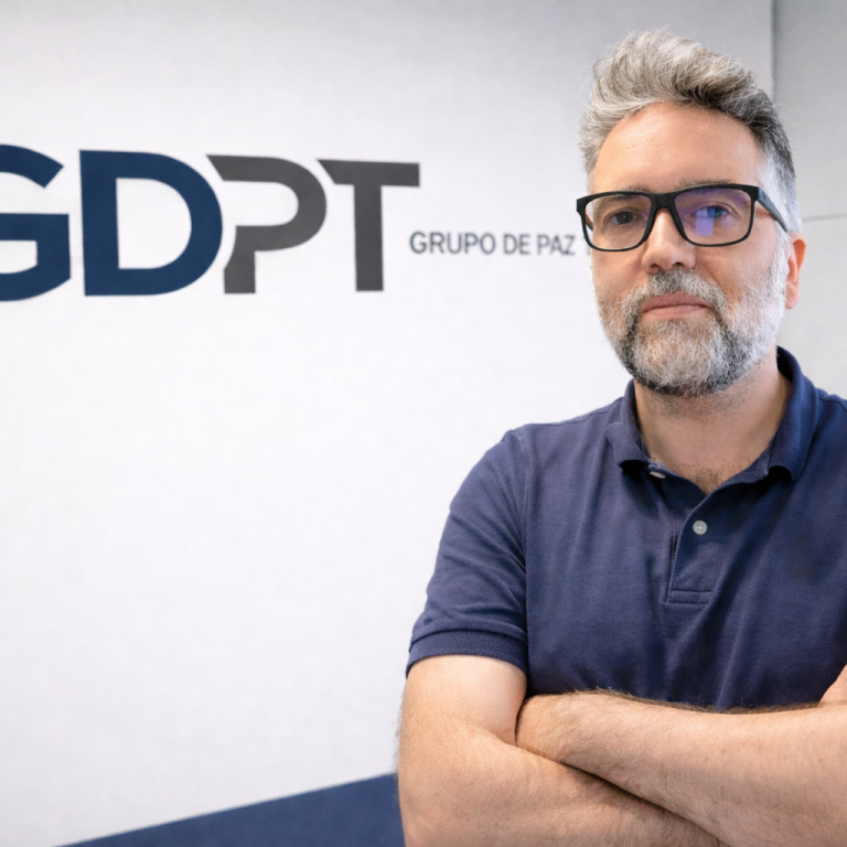 Hombre con gafas y cabello gris, de brazos cruzados, de pie junto a un logo.